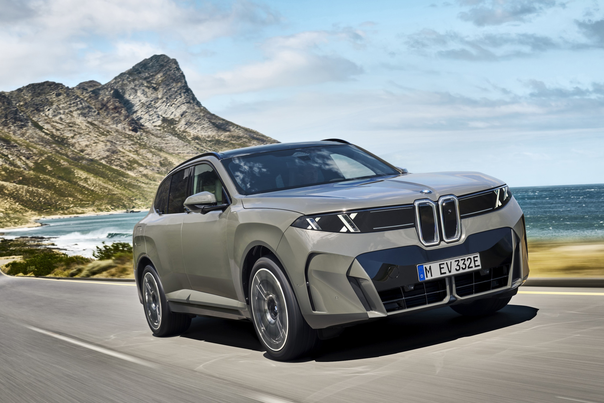 BMW iX5 in Grau fährt auf einer Küstenstrasse vor Bergpanorama – derzeit grösste Batterie aller Elektro-PKW.