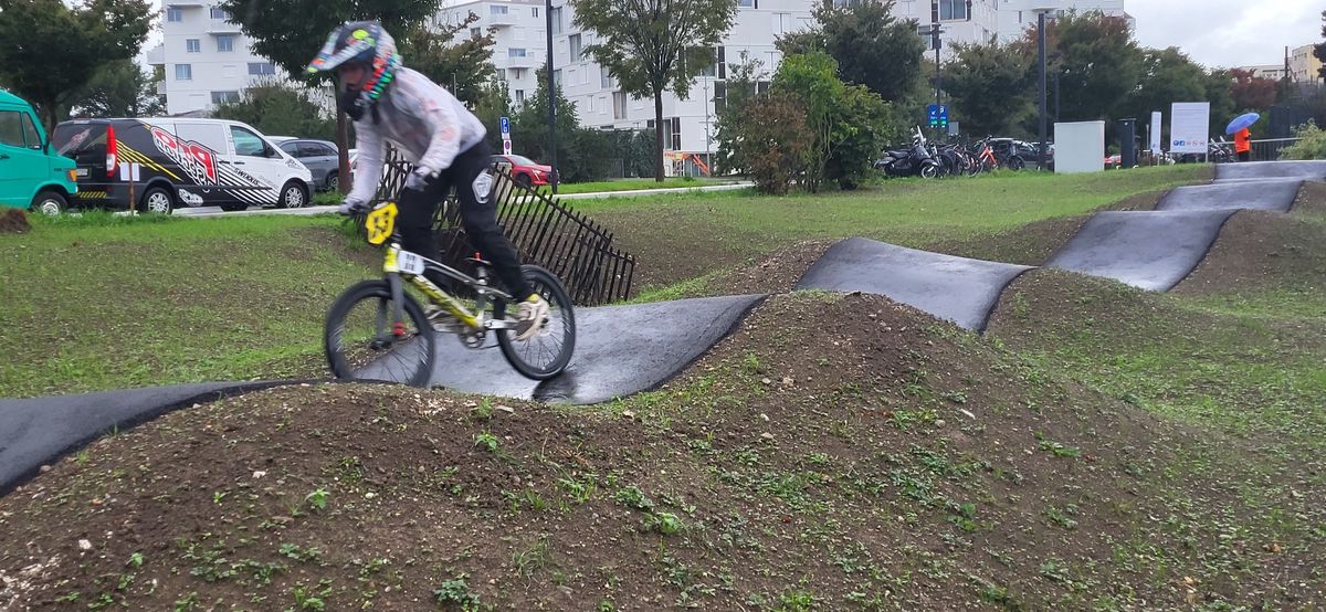 Cycliste en BMX sur une piste de bosses en plein air avec des immeubles en arrière-plan.