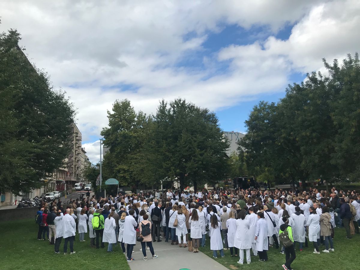 Groupe de personnes en blouses blanches se rassemblant dans un parc, entouré de bâtiments et d'arbres par une journée nuageuse.