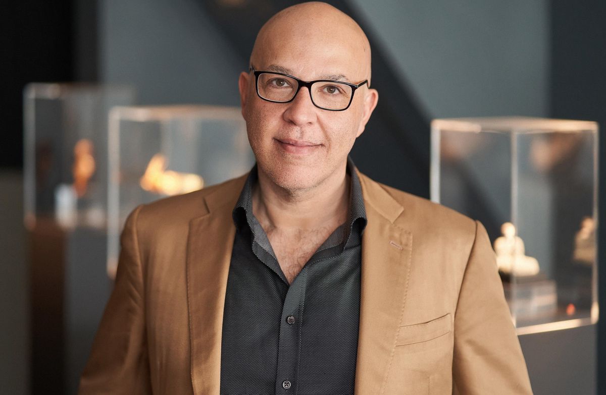 Homme en veste beige et chemise noire devant des vitrines d’exposition.
