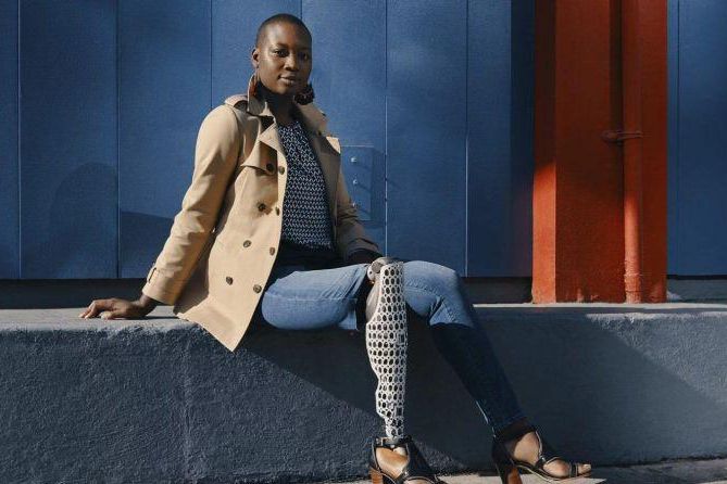 Personne assise sur un banc, portant un trench-coat beige et un pantalon avec un motif unique, devant un mur bleu.