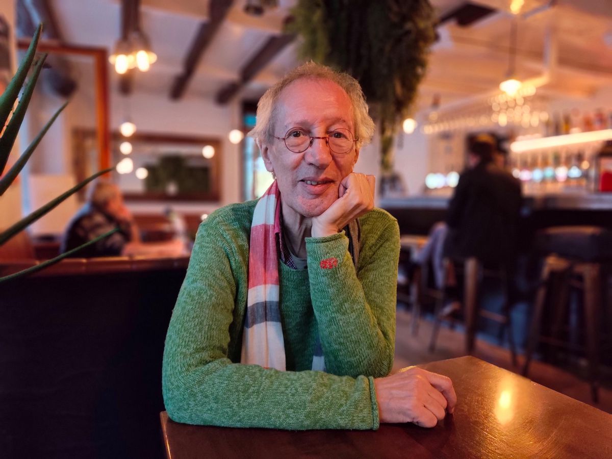 Personne âgée portant un pull vert et une écharpe assise à une table dans un café avec un éclairage chaleureux.