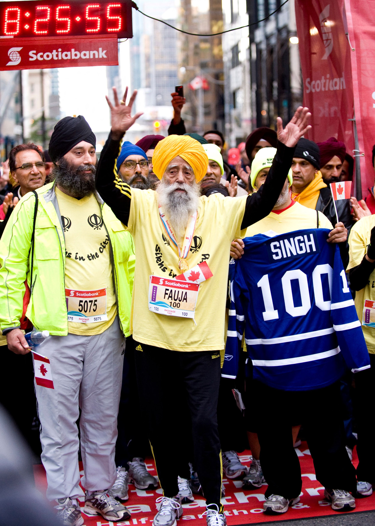 Der 100-jährige britische Sikh Fauja Singh beendet den Scotiabank Toronto Waterfront Marathon 2011 und hebt triumphierend die Arme, umgeben von Unterstützern in gelben Shirts.