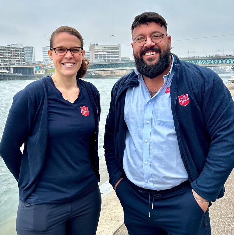 Zwei Personen in Freizeitkleidung mit Abzeichen der Heilsarmee stehen lächelnd an einem Flussufer. Im Hintergrund sind Gebäude und eine Brücke zu sehen.