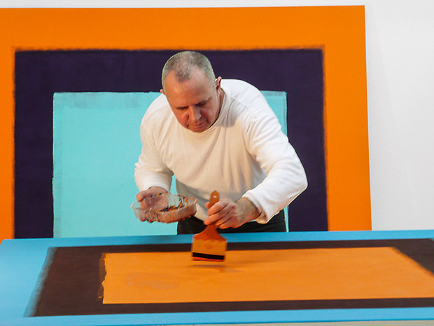 Ein Künstler malt ein geometrisches Bild mit kräftigen Farben wie Orange, Blau und Schwarz.