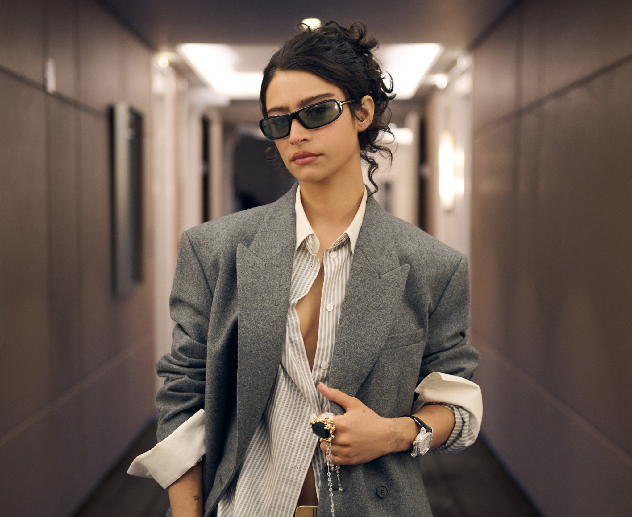 Stylish young woman wearing an oversized gray blazer, striped shirt and black sunglasses in a cozy hallway.