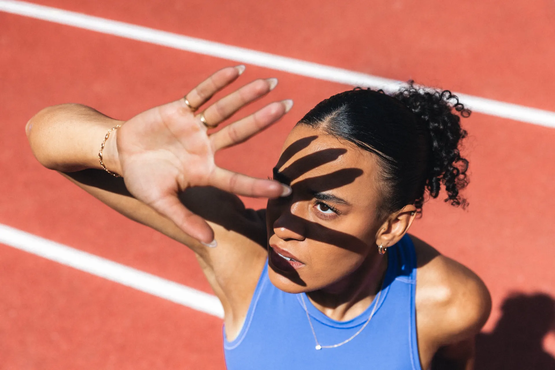 Ditaji Kambundji auf roter Laufbahn hält Hand gegen die Sonne, Fingerschatten fällt auf ihr Gesicht, blaues Sporttop.