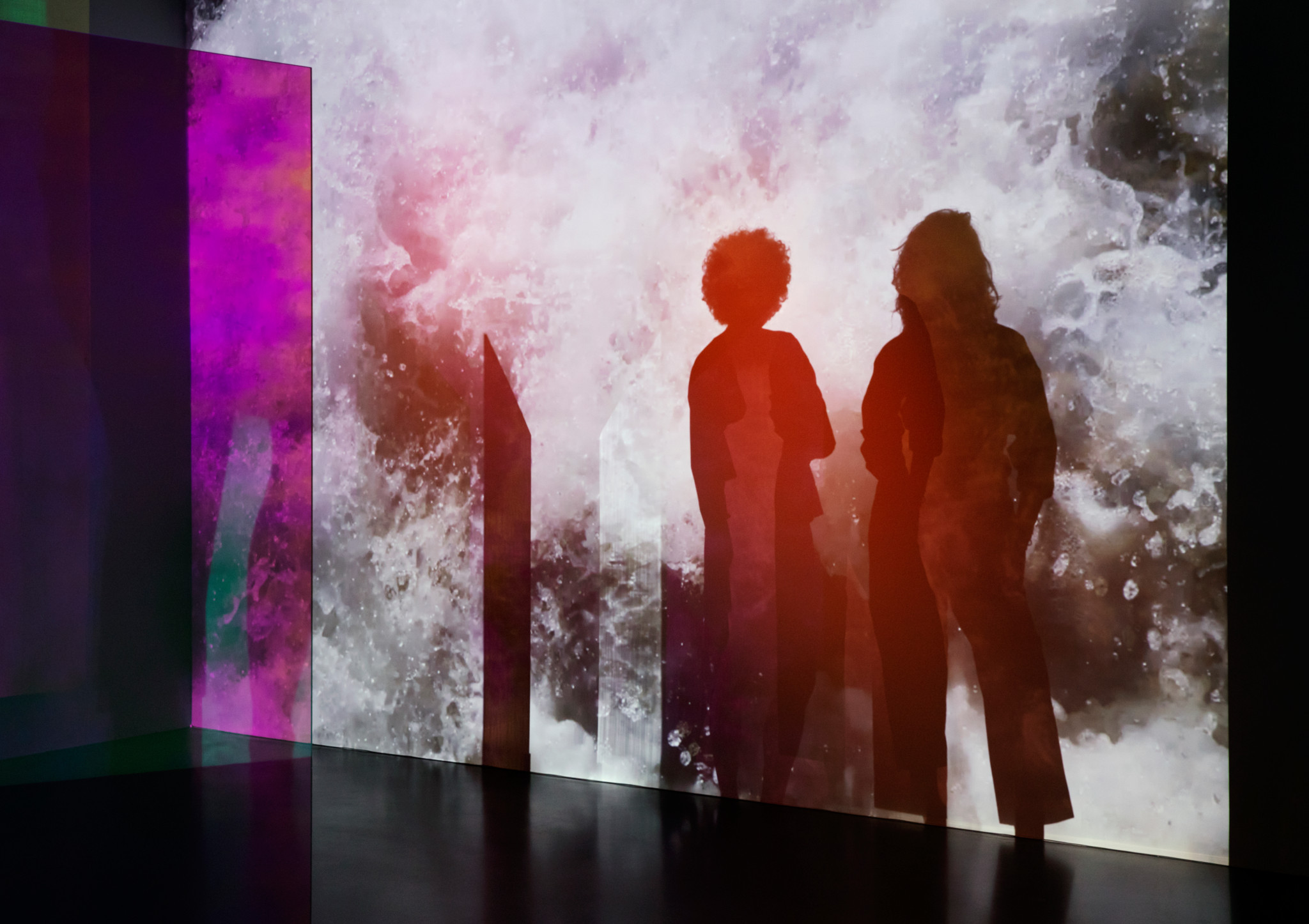 Zwei Personen als Silhouetten vor einer leuchtenden Projektion mit rosa-weissen Farben und bunten Glasscheiben in einer Kunstinstallation.