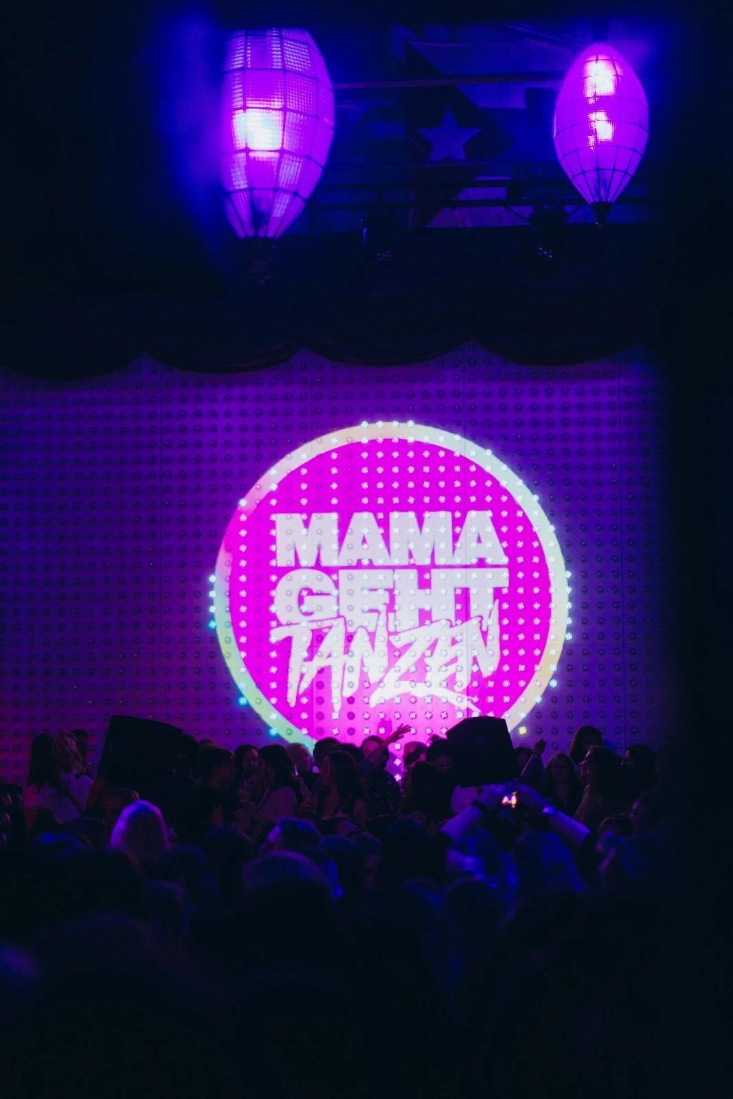 Leuchtende Projektion mit Text ’Mama geht tanzen’ auf einer Party. Personen tanzen im Vordergrund unter bunten Lichtern.