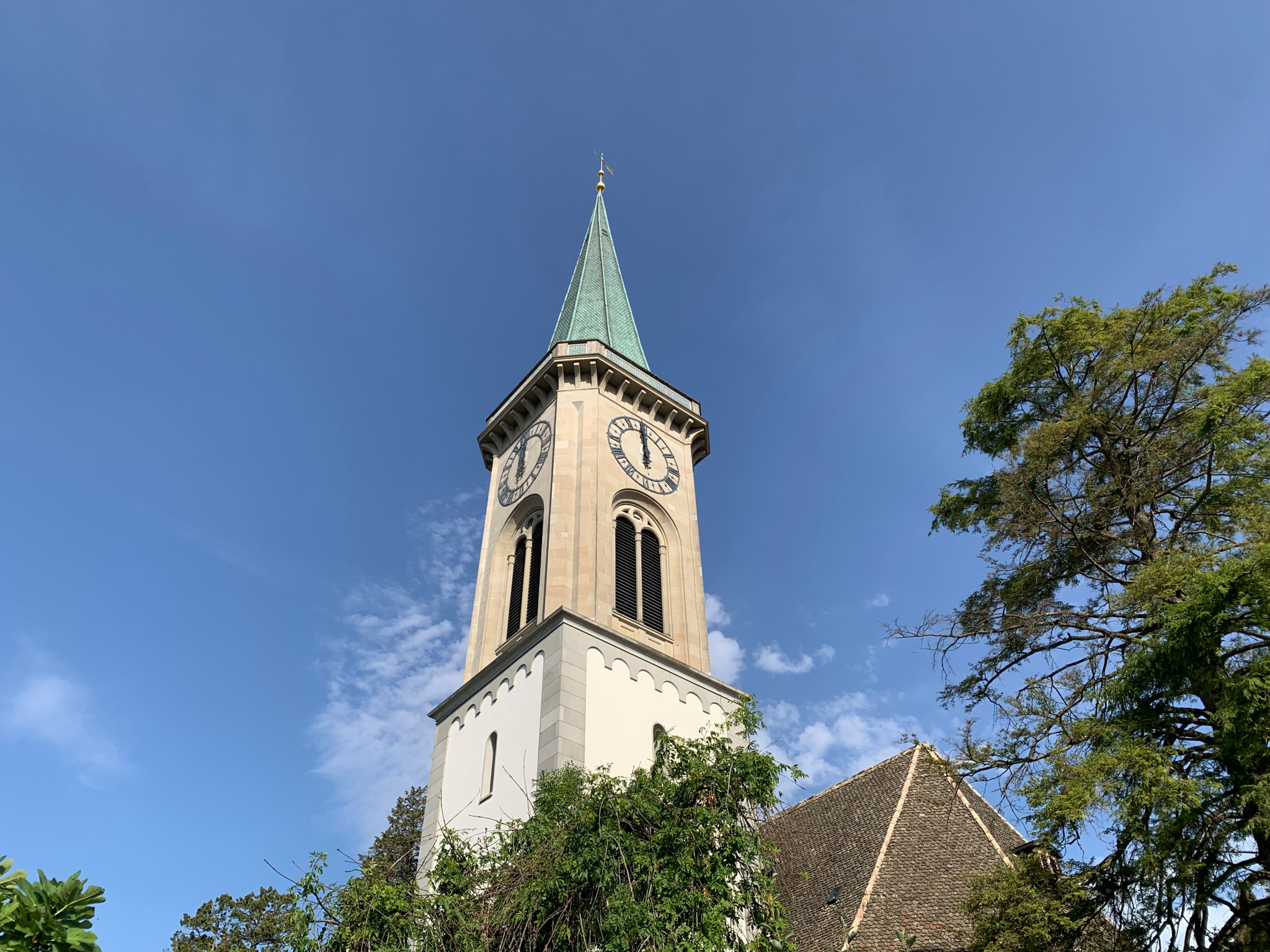 Kirchturm mit grünem Spitzdach zwischen Bäumen, im Hintergrund der Zürichsee unter blauem Himmel.