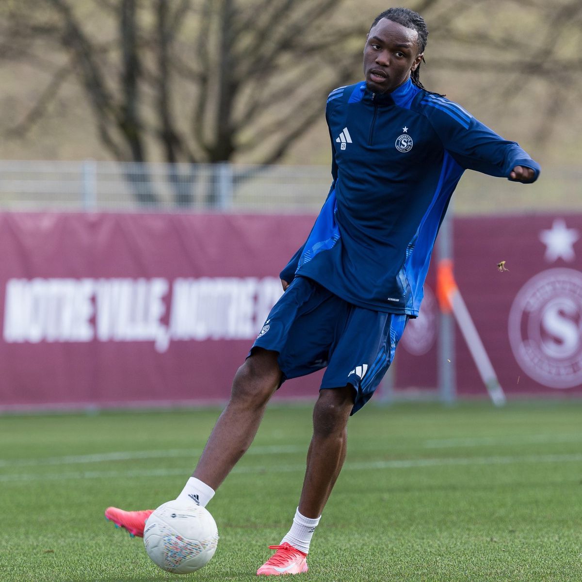 Un joueur de football du Servette FC, Joseph Nonge, en tenue d’entraînement bleue effectue une passe sur le terrain.