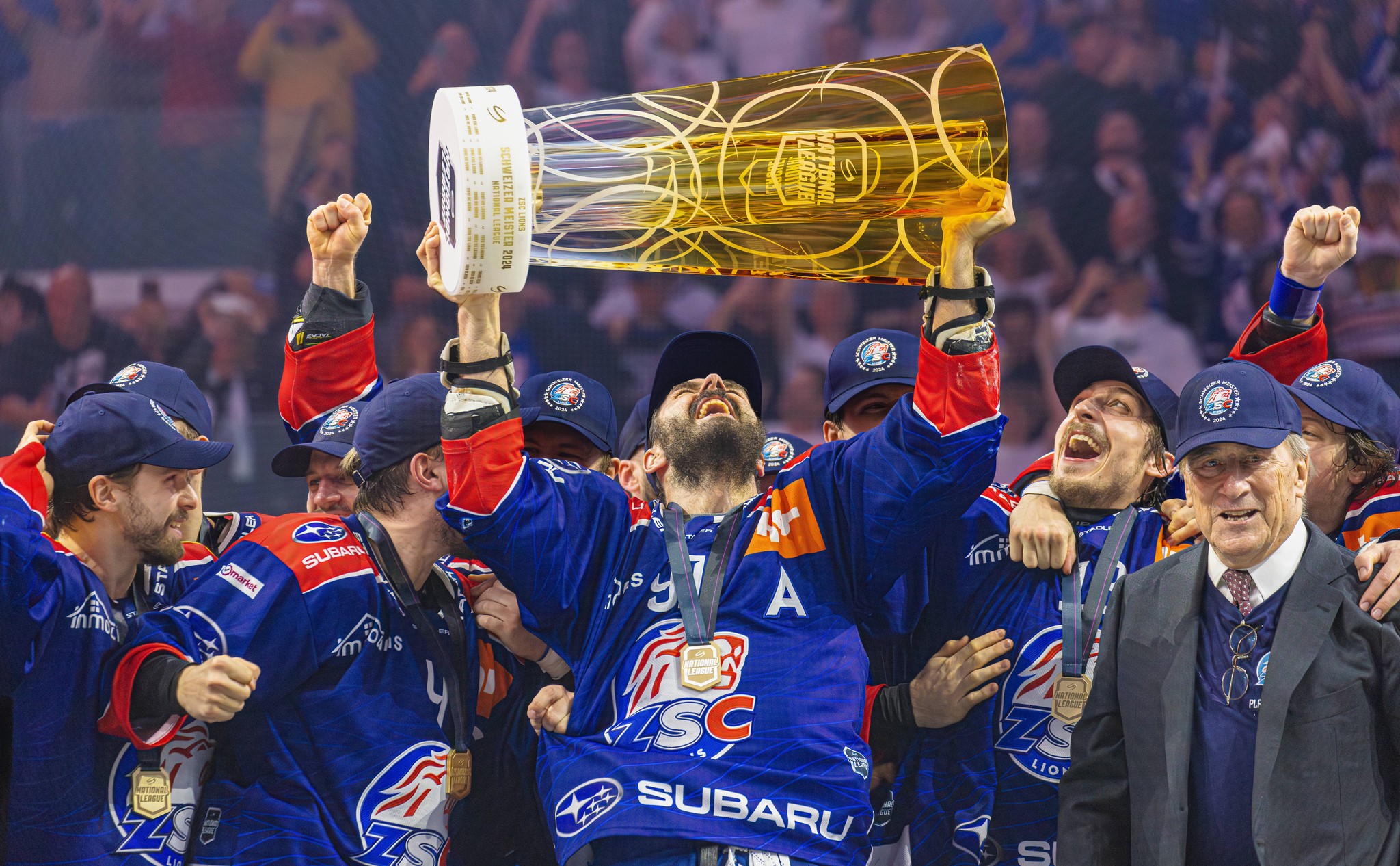 ZSC Lions-Stürmer Denis Hollenstein hebt den Pokal nach dem Gewinn des Schweizer Meistertitels 2023/2024 in Zürich, umgeben von jubelnden Teamkollegen.