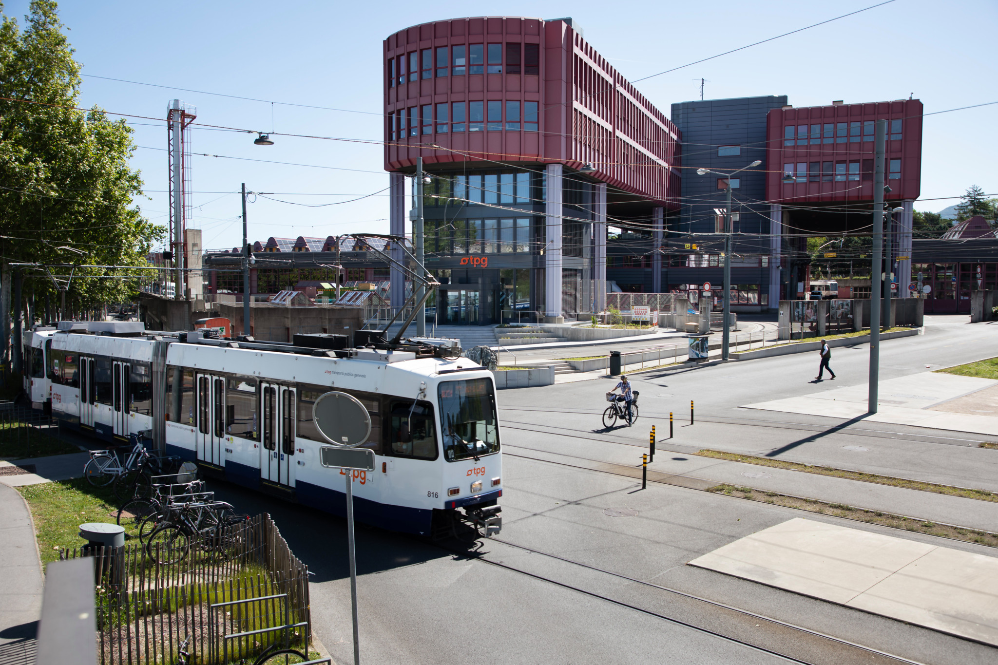Vue du site du Bachet-de-Pesay à Genève, abritant le siège des tpg avec un tramway passant devant le bâtiment moderne. Un cycliste et un piéton traversent la route.