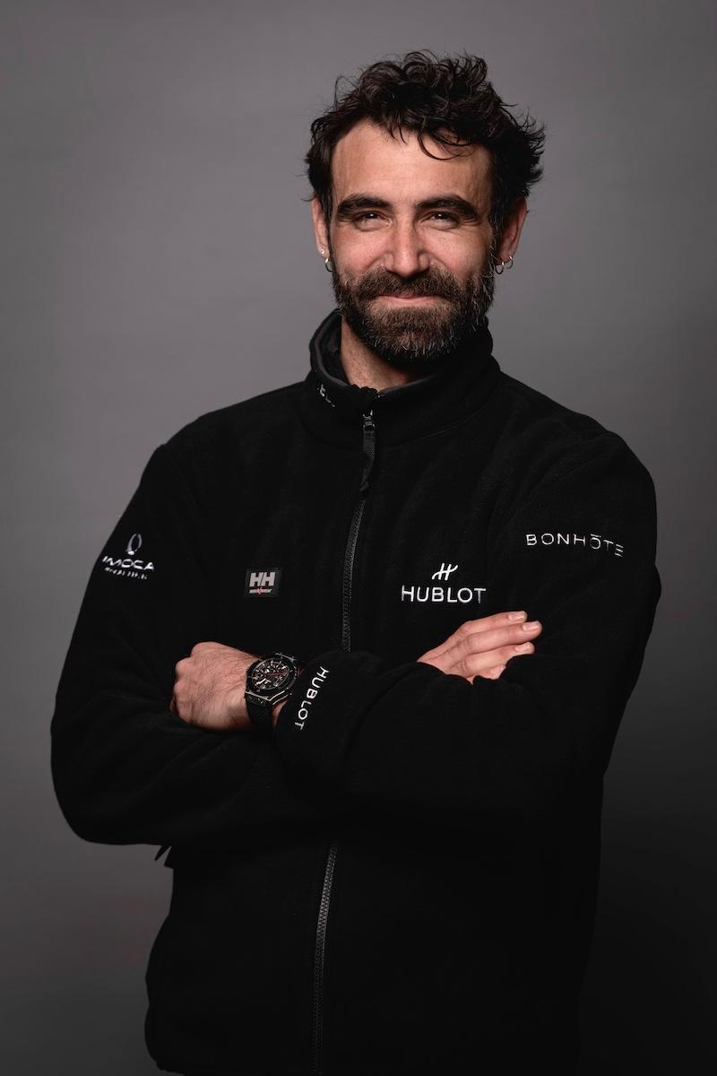 Homme portant une veste noire avec des logos de marques, bras croisés, sur un fond gris.
