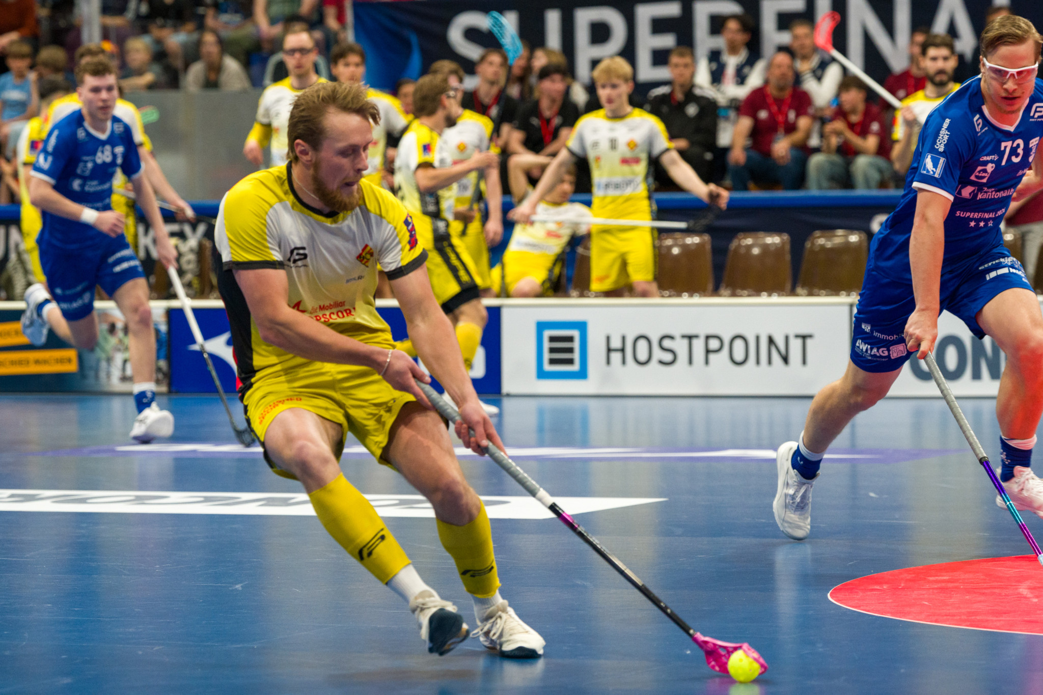 Unihockeyspieler im gelben Trikot schiesst den Ball beim Superfinal, verfolgt von einem Gegner in Blau mit Nummer 73.