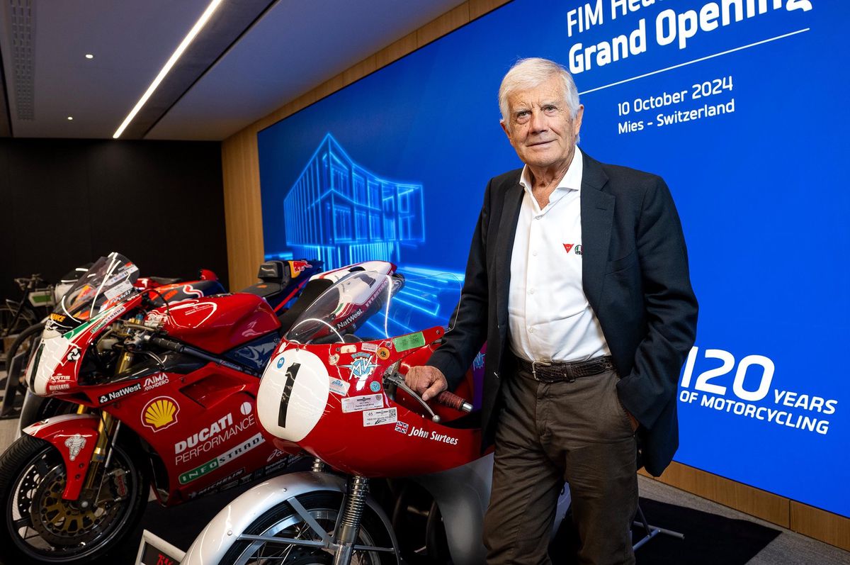 Giacomo Agostini lors de l'inauguration du nouveau siège de la Fédération internationale motocycliste à Mies, posant devant des motos, avec un panneau indiquant l'ouverture des nouveaux bureaux de la FIM en arrière-plan.
