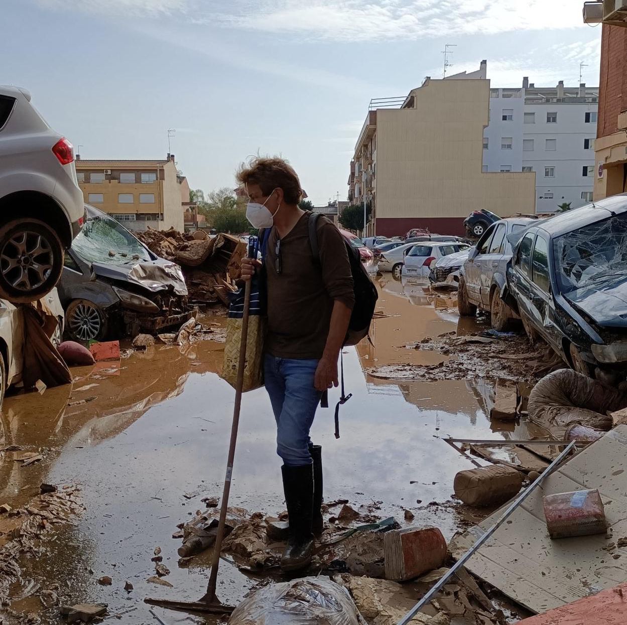 Homme avec un masque et un bâton marchant dans une rue inondée à Valence, avec des voitures endommagées et des débris autour.