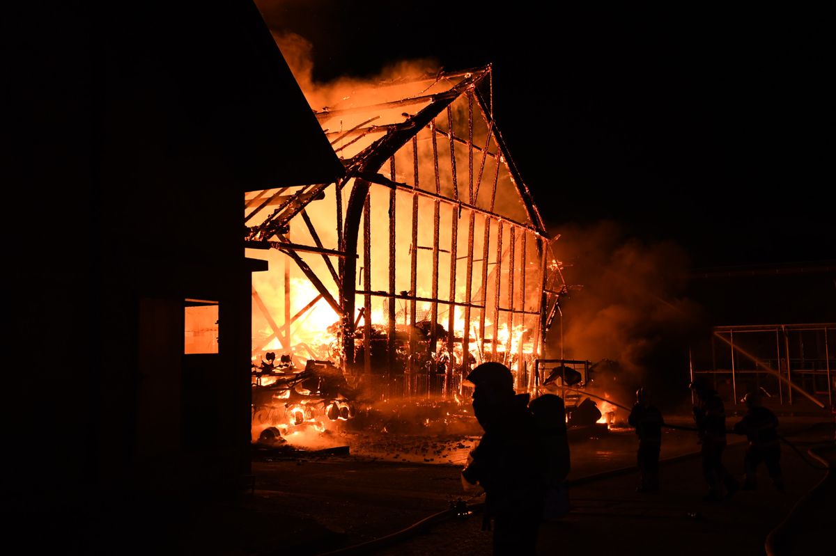 Un bâtiment en bois en feu la nuit, avec des pompiers en silhouette essayant de contrôler l'incendie.