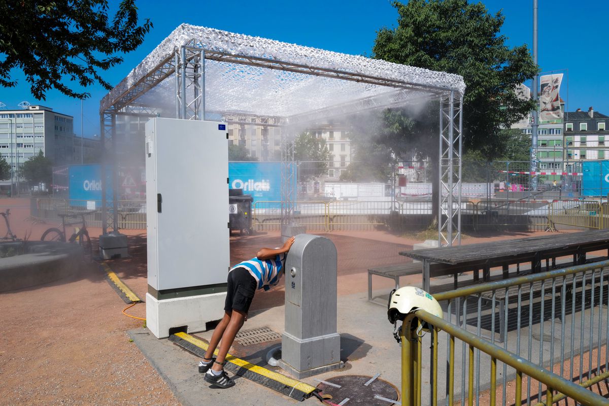 Installation de brumisateurs estivale à Genève avec de grandes structures métalliques sur quatre pieds, un enfant boit à la fontaine devant.