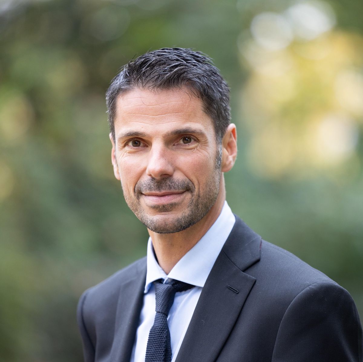 Un homme en costume sombre, portant une chemise blanche et une cravate, sourit devant un fond flou de verdure.