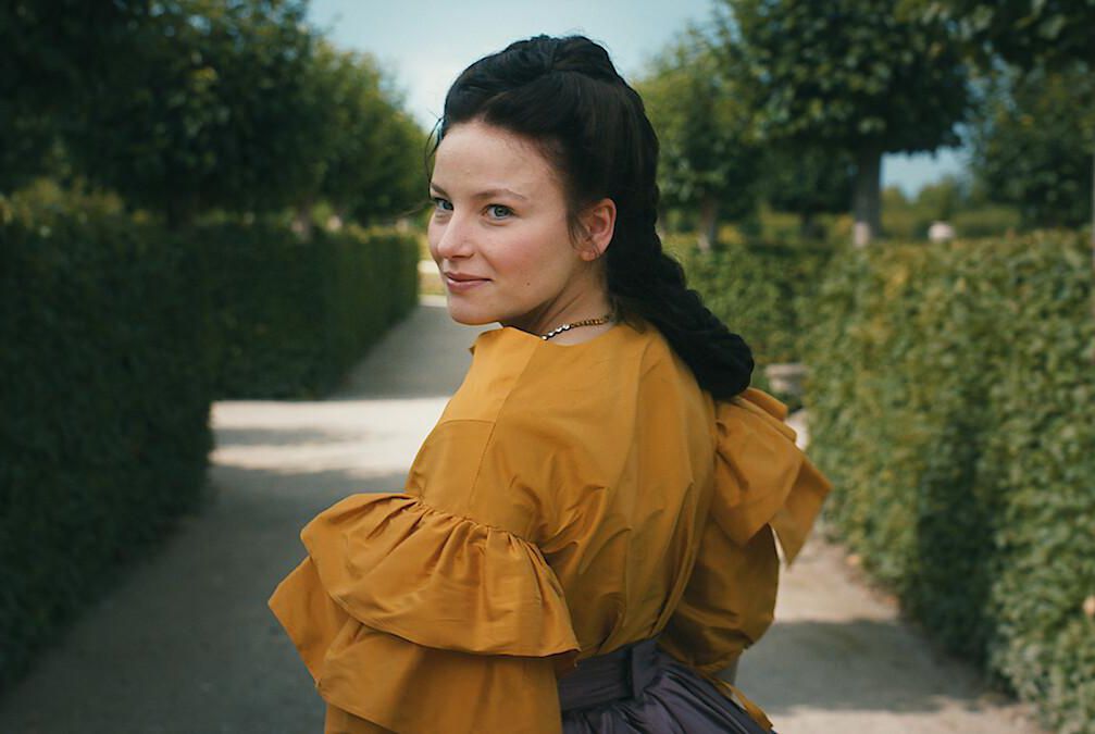 Femme portant une robe jaune dans un jardin entouré de haies, regardant par-dessus son épaule.