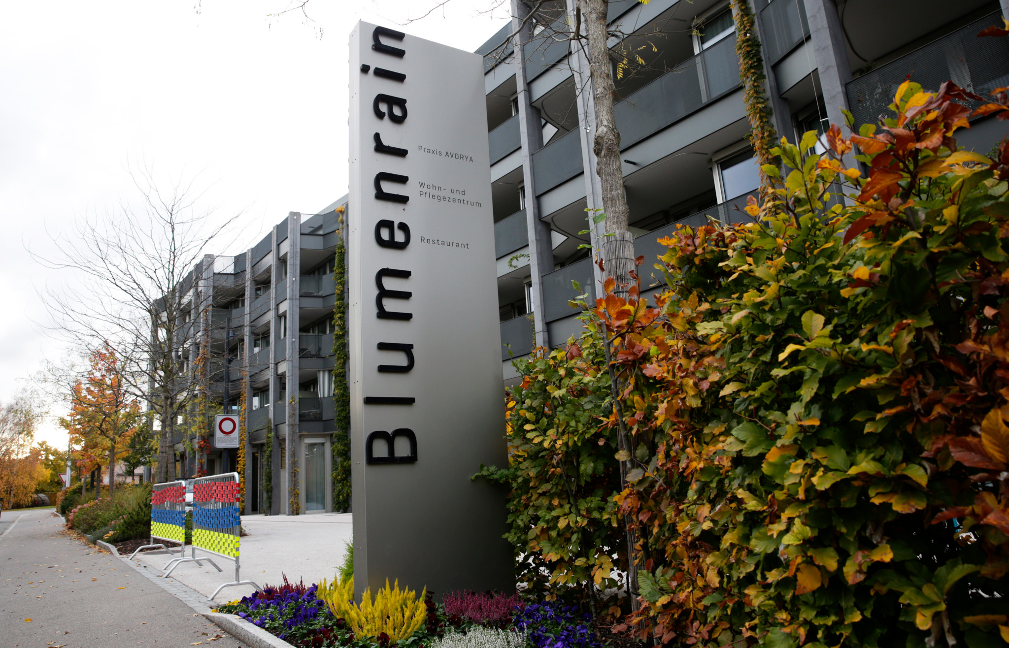 Eingangsschild des Alterszentrums Blumenrain in Zollikon, umgeben von herbstlichem Grün und einem modernen Gebäude im Hintergrund.