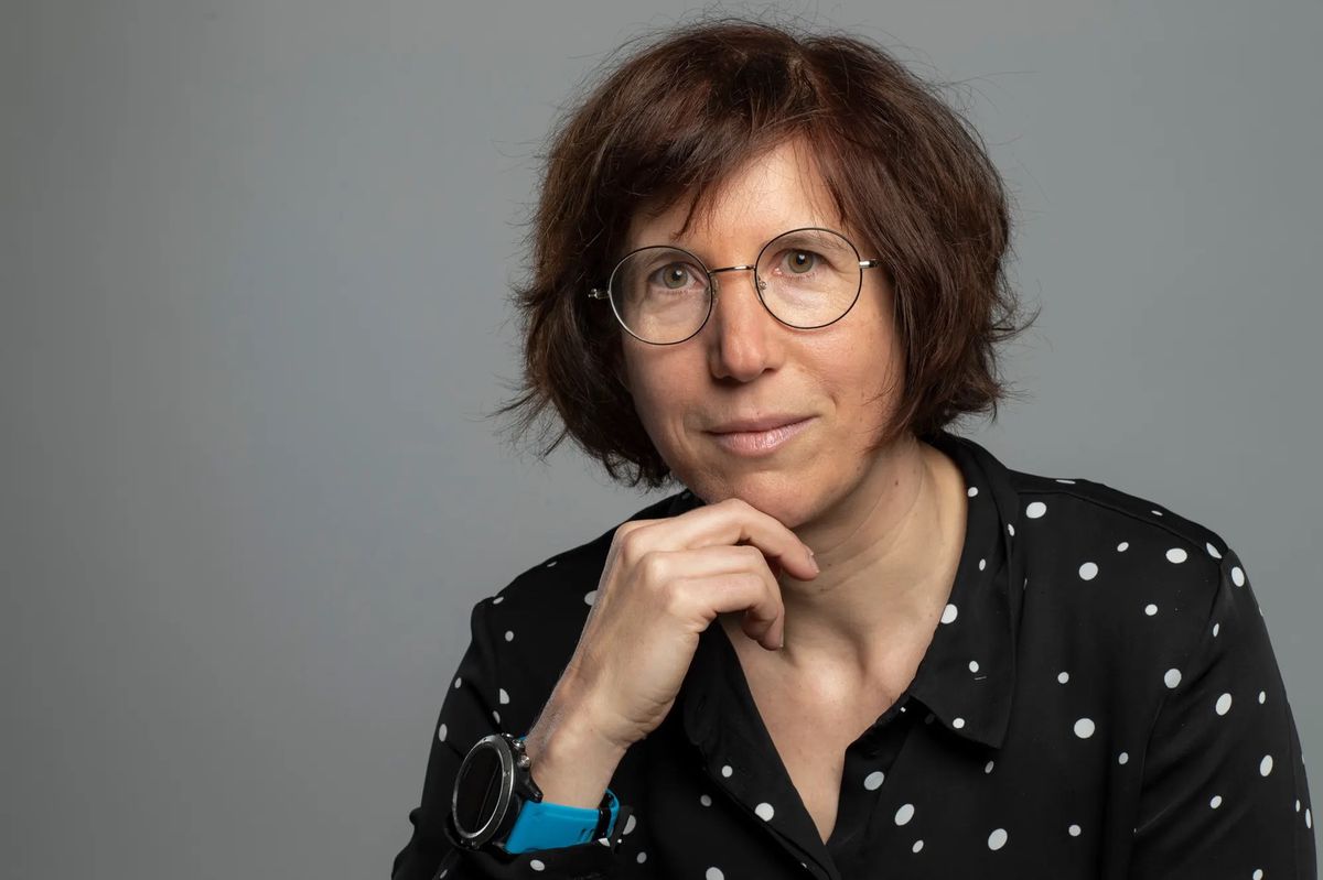 Une femme aux cheveux courts portant des lunettes rondes et une chemise à pois noirs posant avec une main appuyée sous le menton.