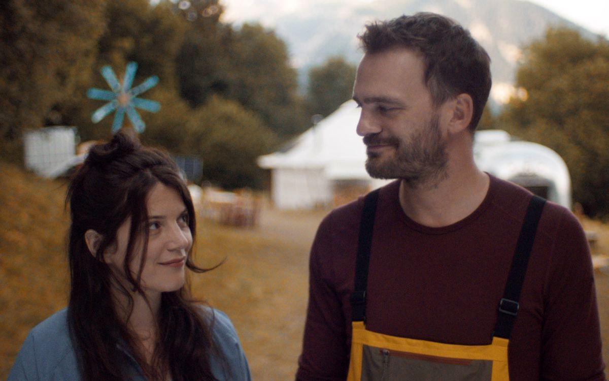 Un homme et une femme marchant ensemble dans un cadre naturel avec des arbres et une tente blanche en arrière-plan.