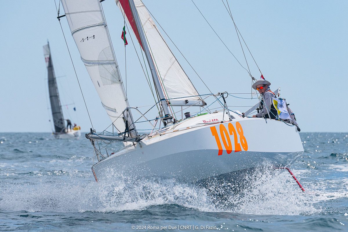 Un voilier blanc avec le numéro 1028 participe à une régate, levant des éclaboussures d'eau alors qu'il navigue. Un autre voilier est visible en arrière-plan.