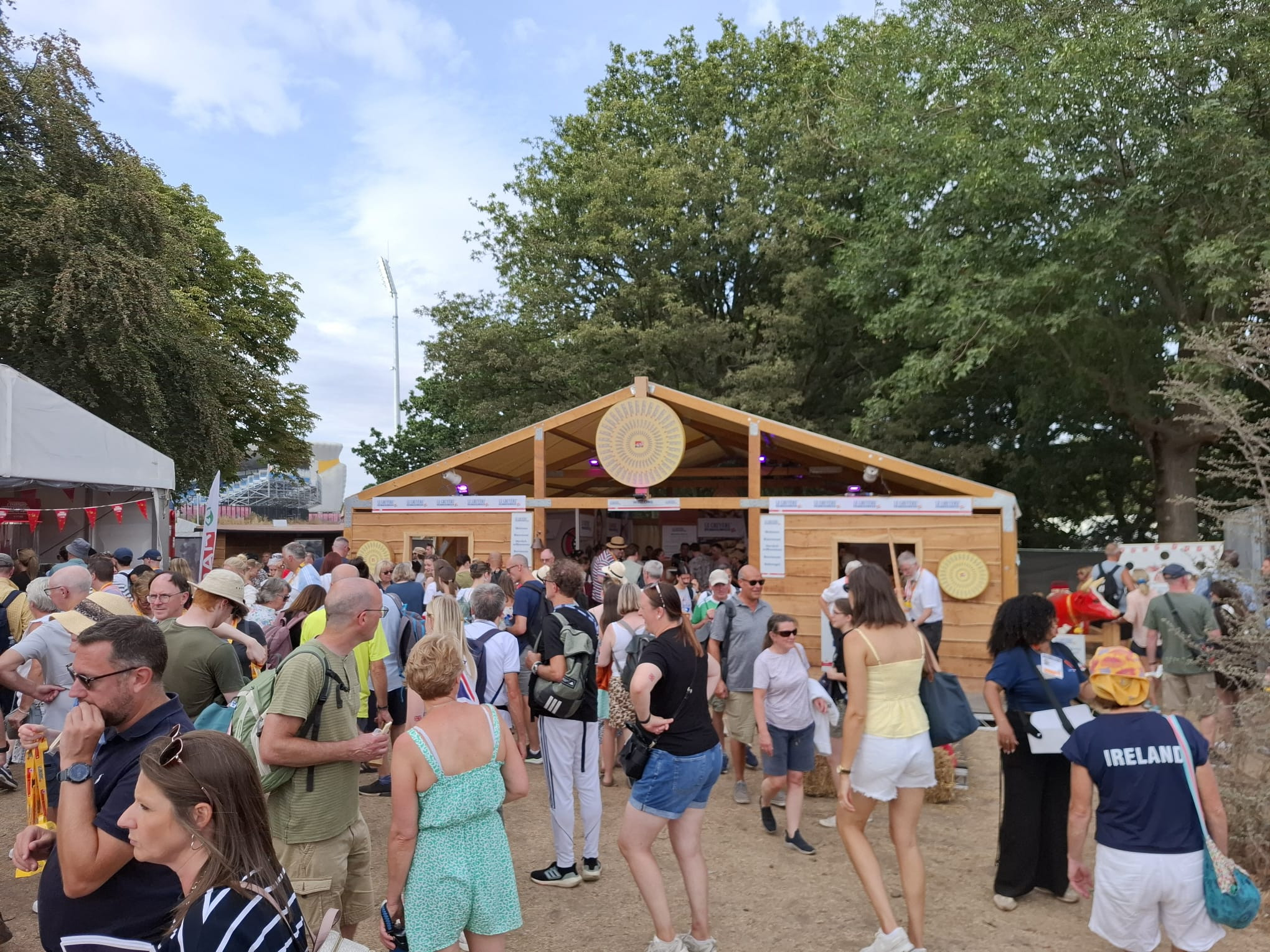 Menschenmenge vor einem Holzpavillon an einem Open-Air-Festival bei sommerlichem Wetter unter grünen Bäumen.