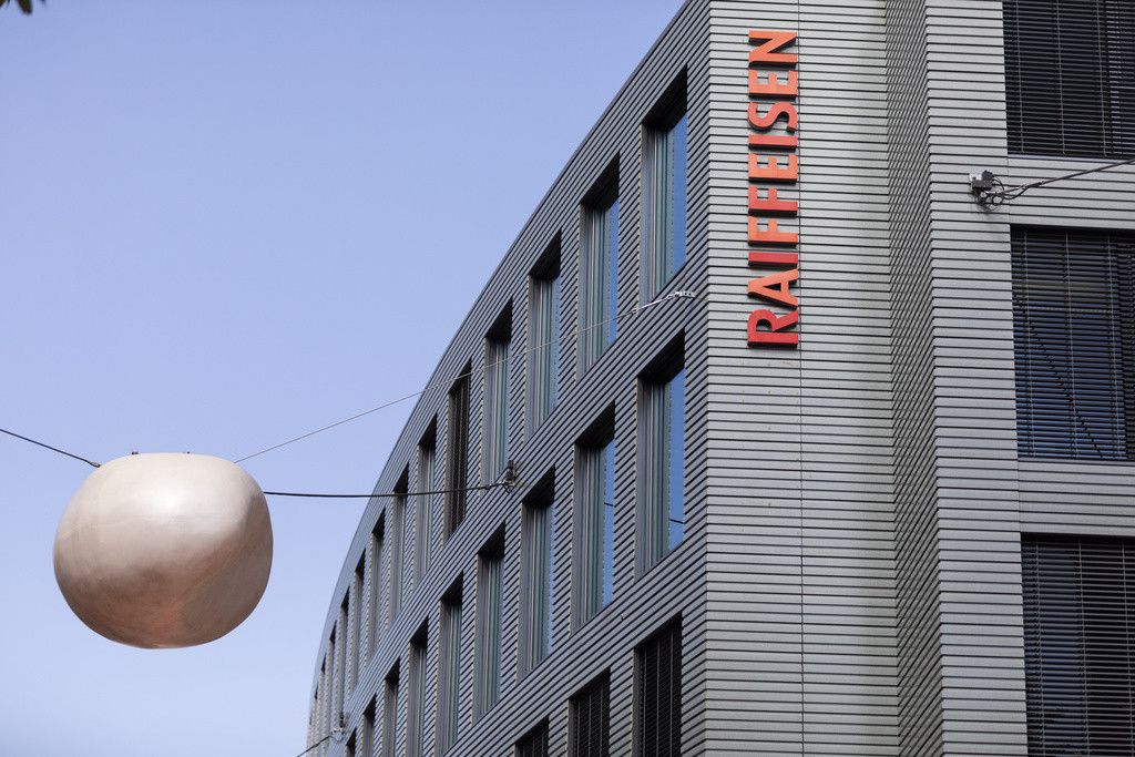 Bâtiment moderne avec des fenêtres alignées et logo Raiffeisen en rouge sur la façade grise, une structure sphérique suspendue au premier plan.