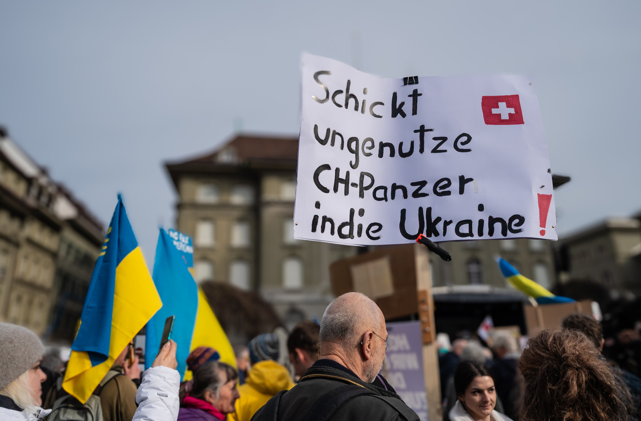 Demonstrant hält Schild mit Aufschrift «Schickt ungenutzte CH-Panzer in die Ukraine» bei Kundgebung mit ukrainischen Flaggen.