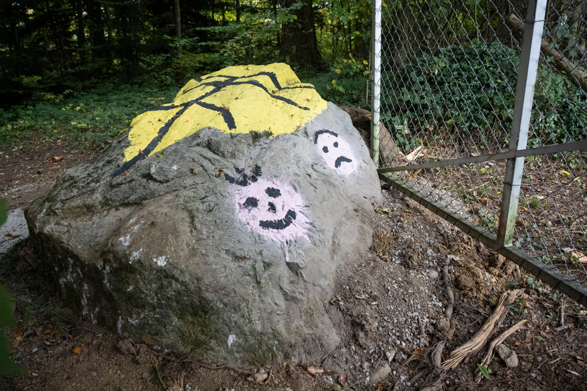Grosser Felsen im Strättligwald, bemalt mit fröhlichen und traurigen Gesichtern neben einem Metallzaun.