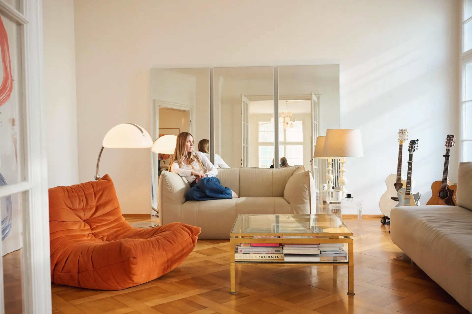 Frau sitzt auf beigem Sofa in hellem Wohnzimmer mit orangenem Sessel, Glastisch und Gitarren an der Wand.