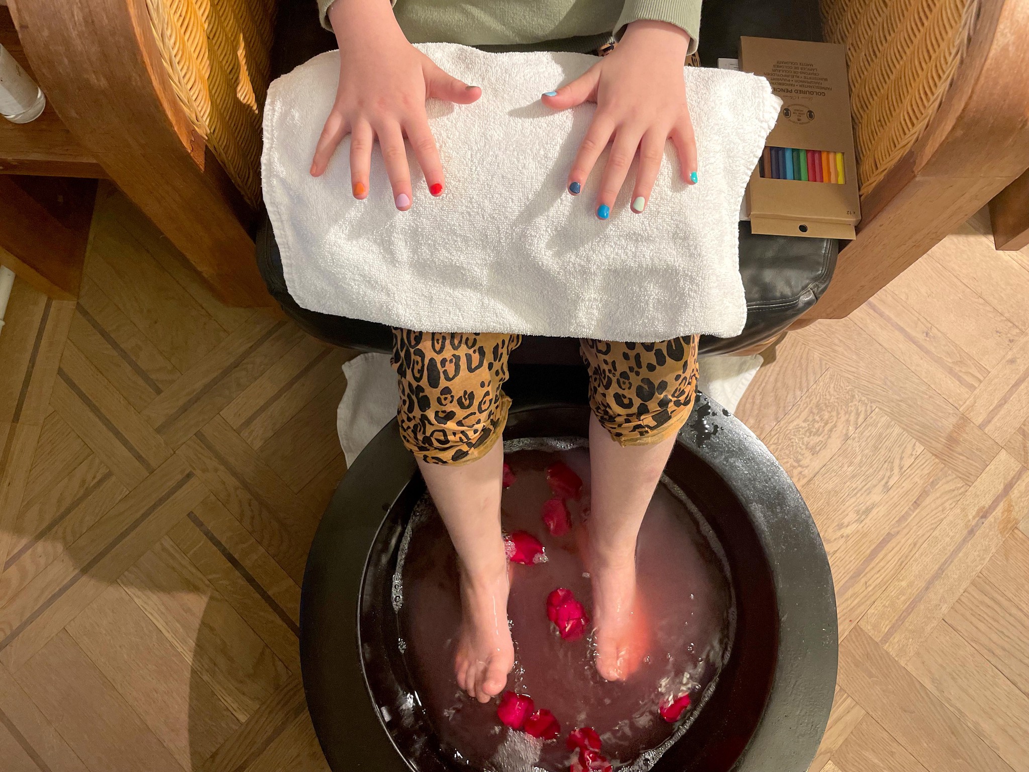Kind macht ein Fussbad in der Schminkbar in Zürich. Es sitzt da, badet seine Füsse im Wasser, das mit Rosenblüten dekoriert ist und hat bunt bemalte Fingernägel.
