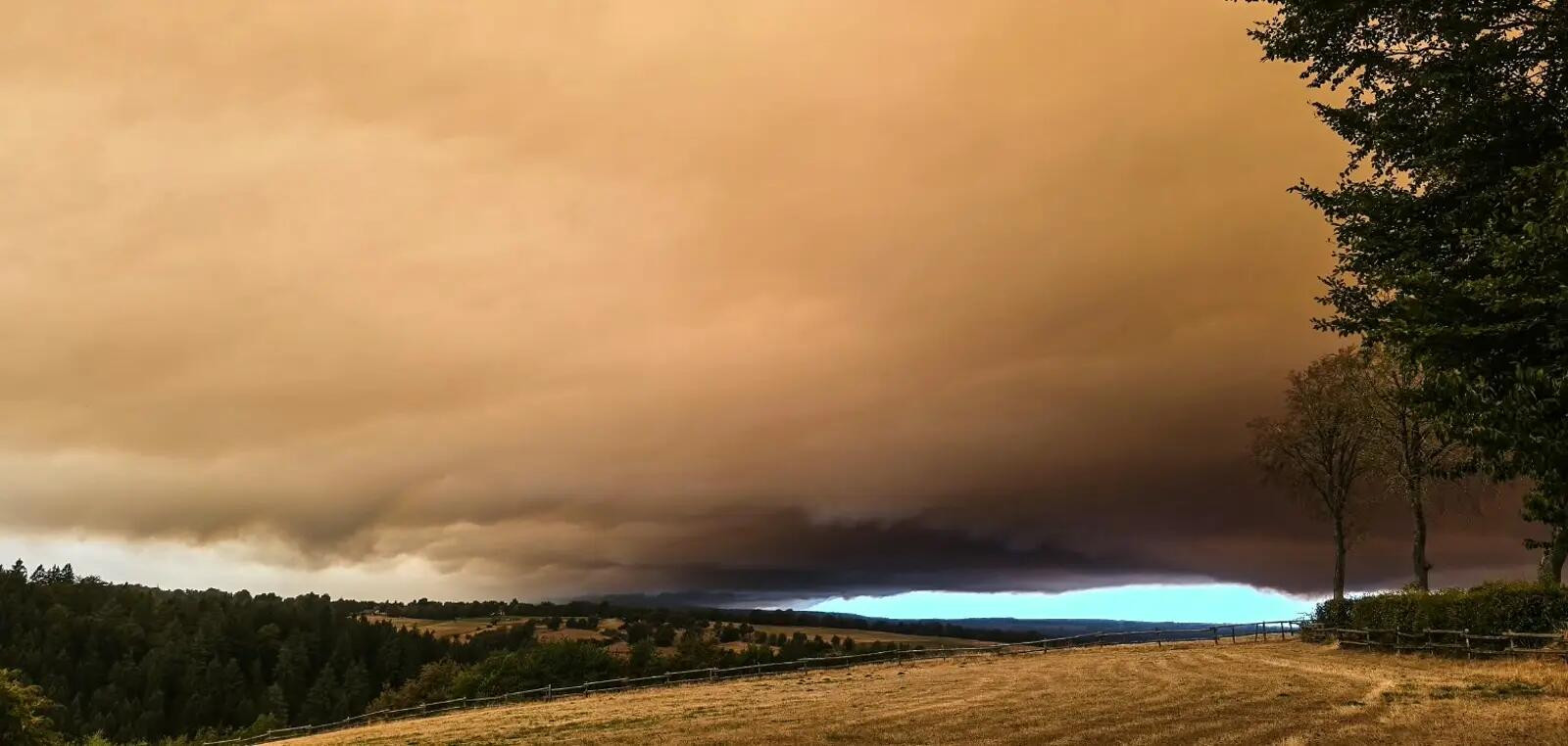 Dramatischer orangefarbener Himmel mit dunklen Rauchwolken über einer ländlichen Landschaft mit Feldern und Wäldern.