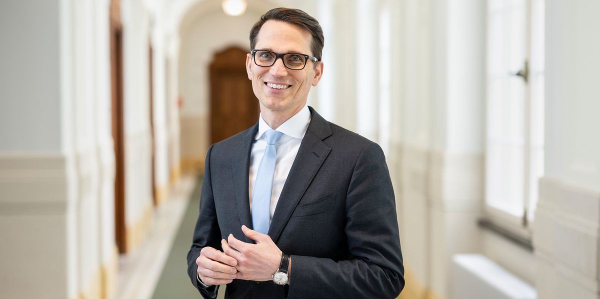 Martin Schlegel in Anzug und Krawatte steht lächelnd in einem hellen, eleganten Flur, umrahmt von Säulen und Türen.