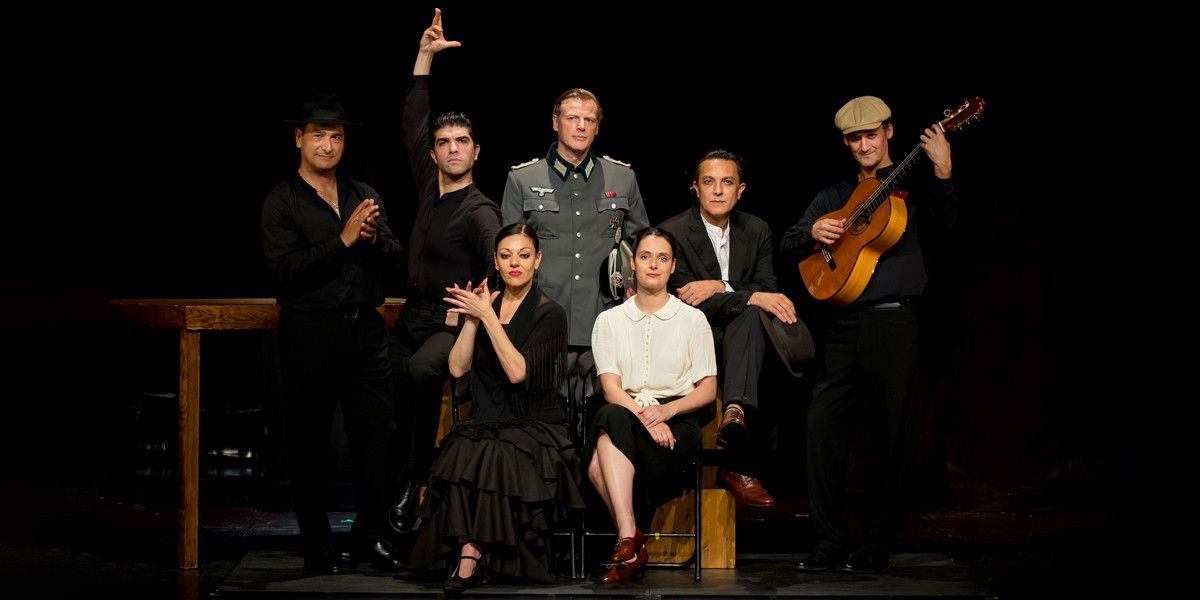 Groupe de sept personnes posant ensemble sur une scène sombre, certains assis et d’autres debout, dont une joue de la guitare.