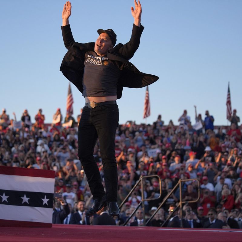 Un homme saute sur scène près d’un orateur à un rassemblement politique avec une foule en arrière-plan.