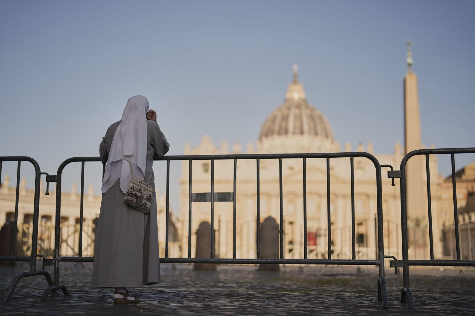 Eine Nonne in grauer Kutte und weissem Schleier betrachtet den Petersdom hinter Absperrungen im Sonnenlicht.