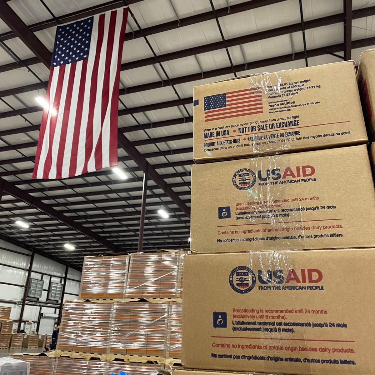 Des cartons de nourriture thérapeutique prête à l’emploi (RUTF) dans l’usine Mana Nutrition à Fitzgerald, Géorgie, prêts à être expédiés, après la réactivation des contrats avec l’USAID. Un drapeau américain est visible en arrière-plan.