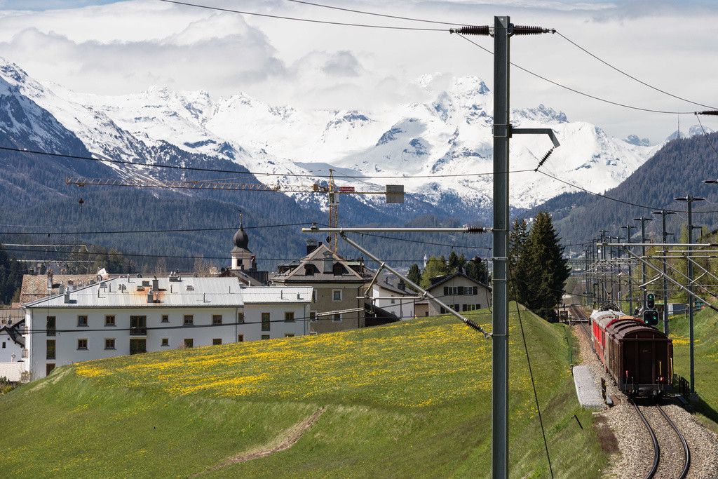 Train rouge près d'un village alpin avec des montagnes enneigées en arrière-plan, champs verts et ciel partiellement nuageux.