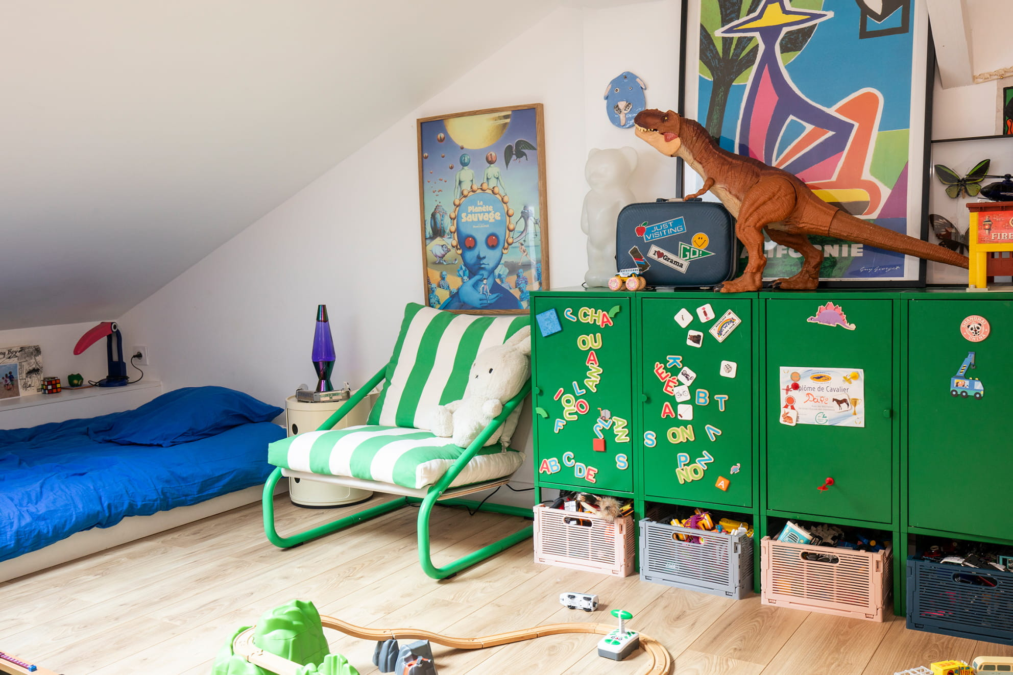 Kinderzimmer mit grünem Metallschrank, Dinosaurier-Figur, bunten Postern, blauem Bett und Spielzeug auf dem Boden