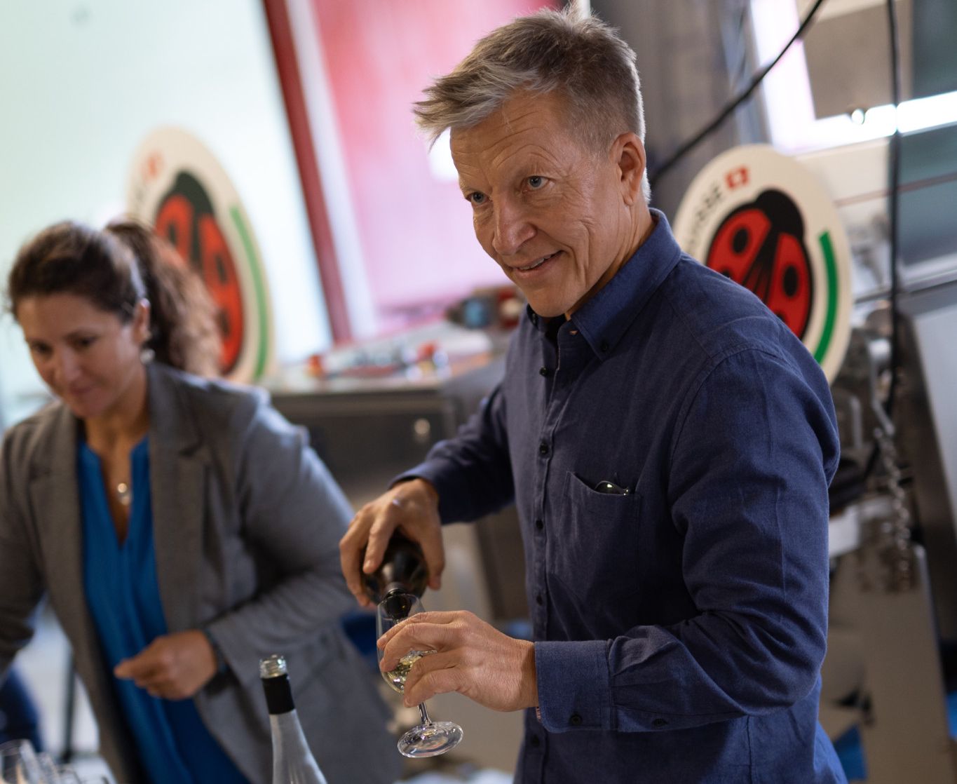 Un homme verse du vin dans un verre sur une table remplie de nombreux verres à vin, avec une femme en arrière-plan.