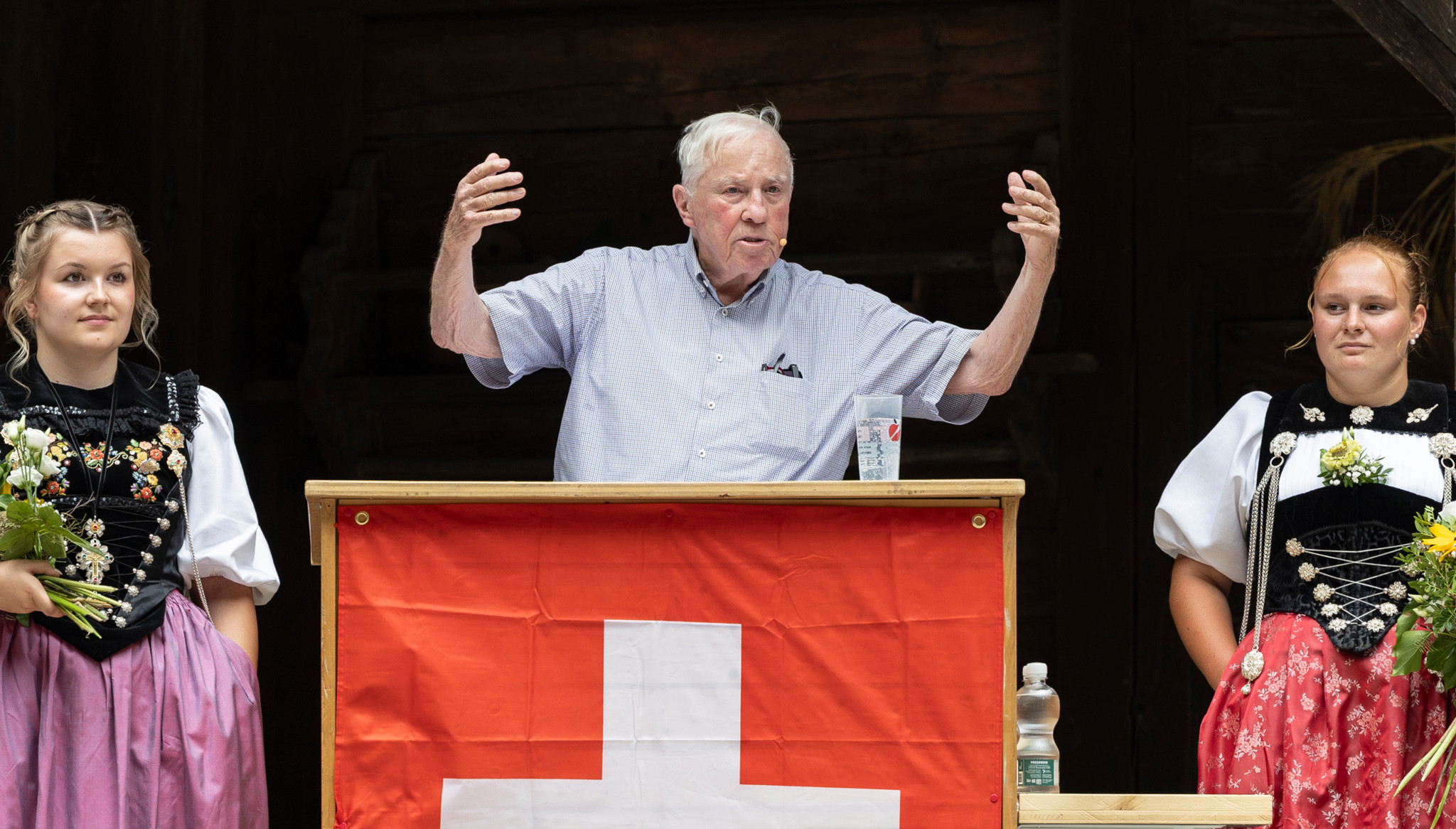 Christoph Blocher hält Bundesfeier-Rede am Tellspiel-Areal Matten bei Interlaken, flankiert von zwei Frauen in Tracht und Schweizer Fahne.