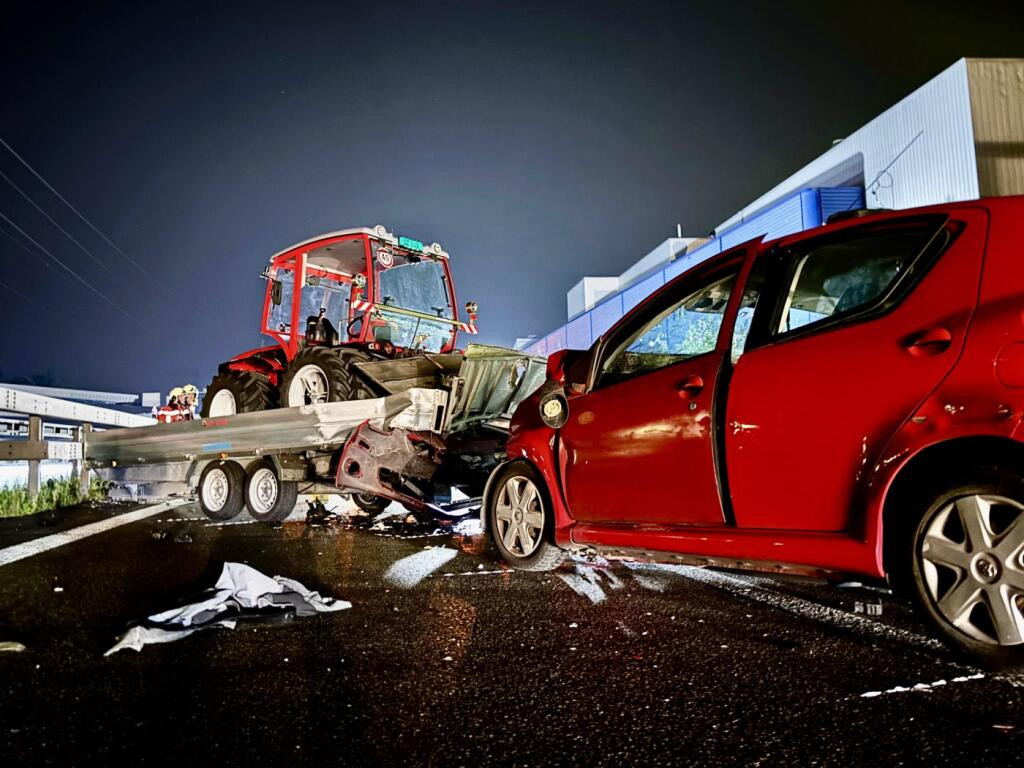 Ein rotes Auto kollidiert mit einem Anhänger und einem Traktor bei Nacht, vor einem Gebäude. Der Unfall geschah nach einer Polizeikontrolle.