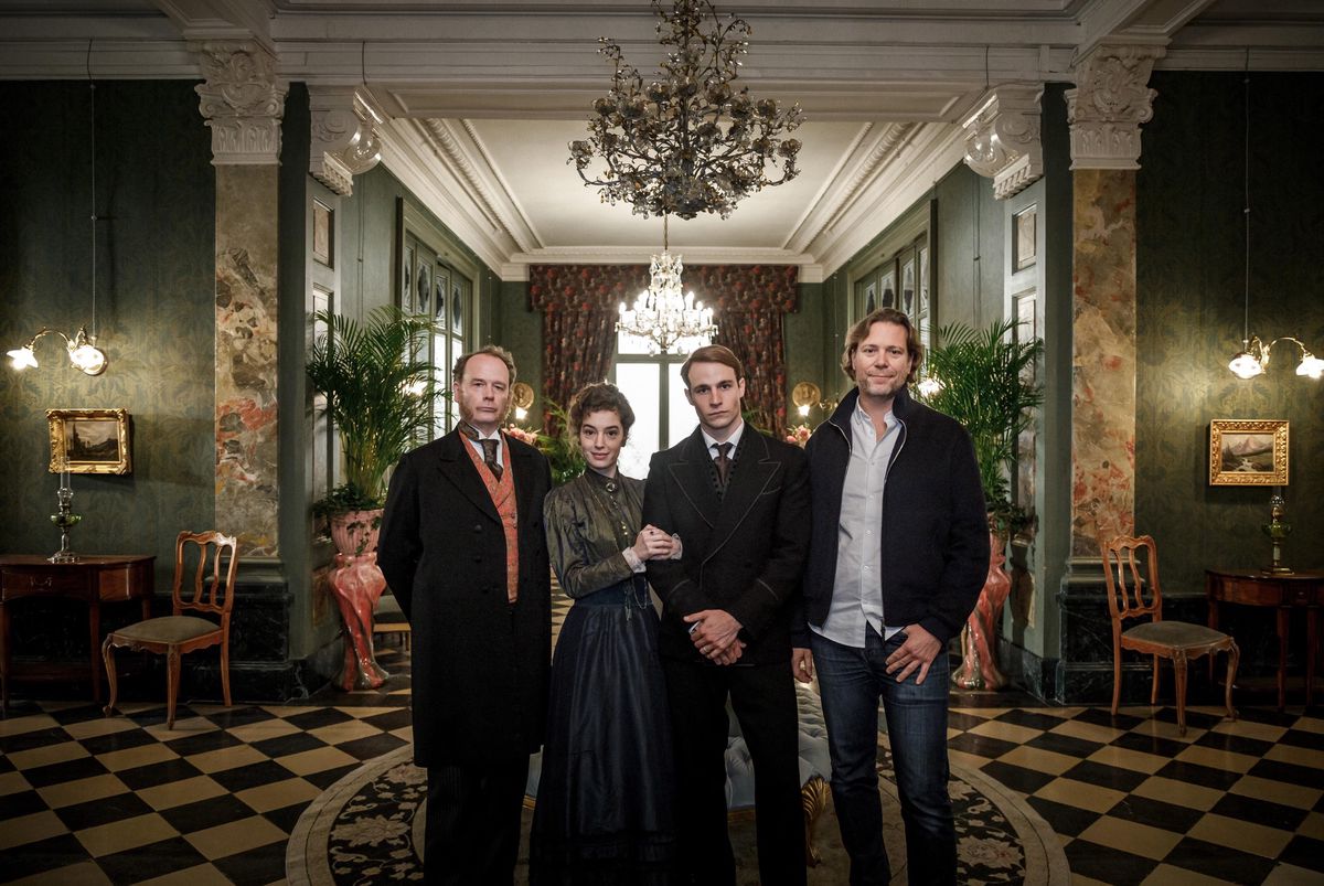 Quatre personnes posent dans un intérieur élégant de style ancien, entouré de lustres et de plantes, pour la série Winter Palace.