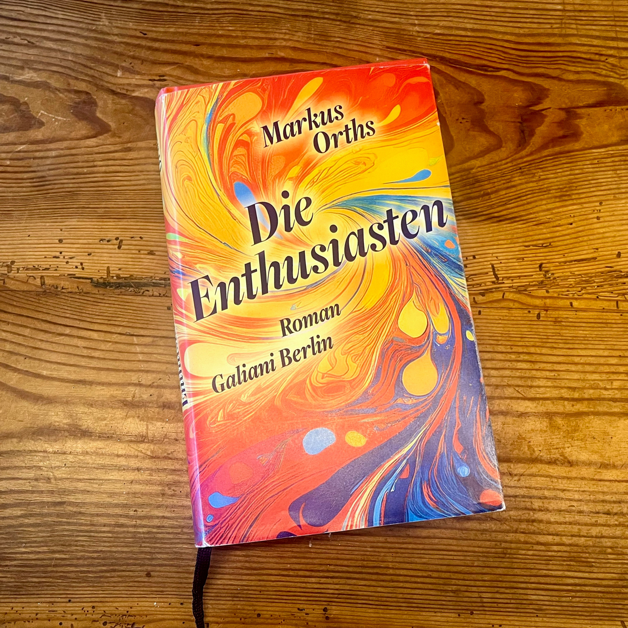 Buchcover ’Die Enthusiasten’ von Markus Orths, Roman im Galiani Berlin Verlag, mit buntem Marmorierungsmuster in Orange, Rot und Blau.