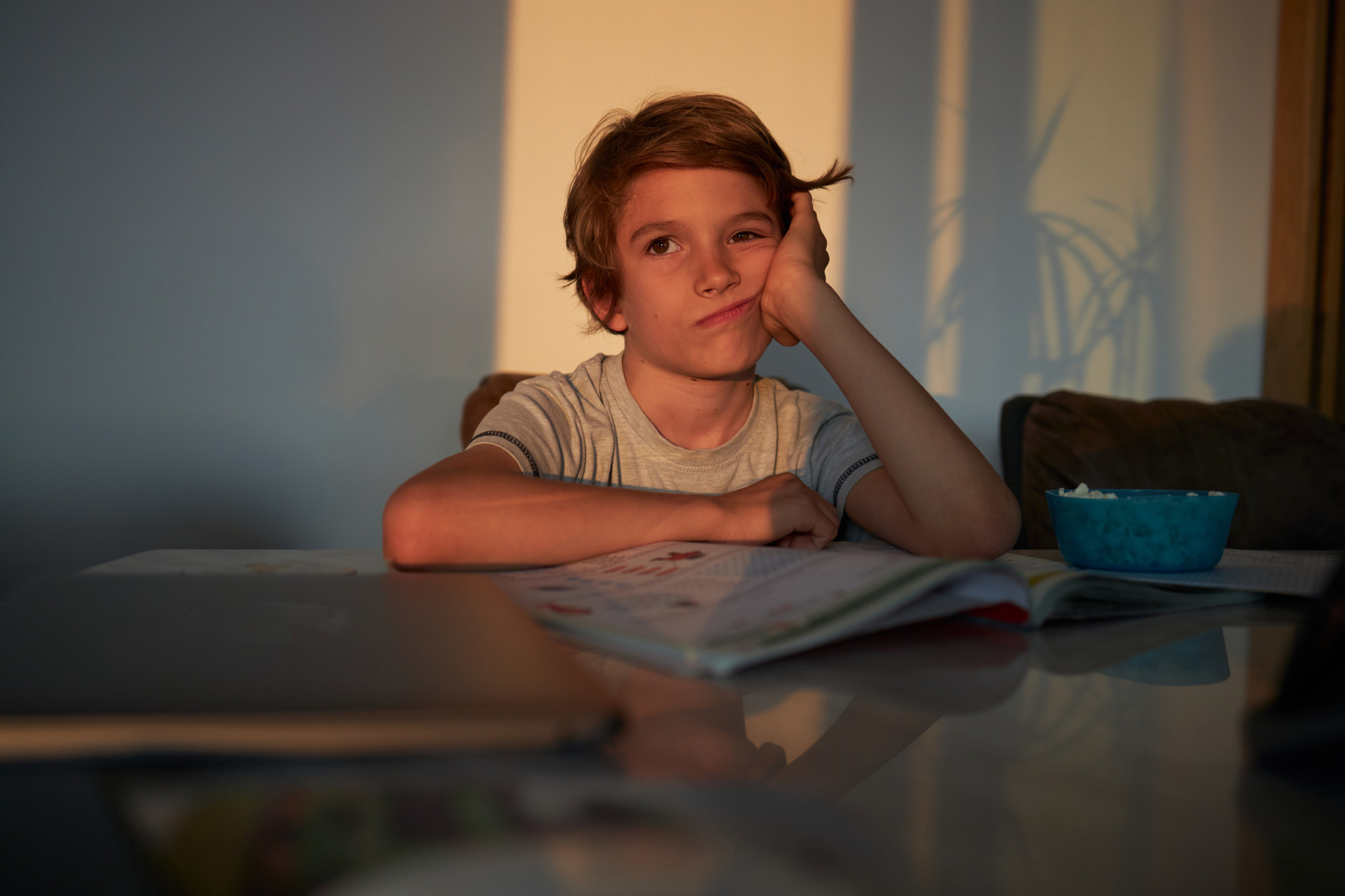 Ein Schuljunge sitzt gelangweilt am Tisch und studiert zu Hause, mit einem aufgeschlagenen Buch vor sich.