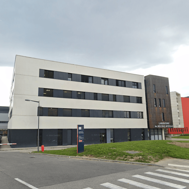 Bâtiment moderne en béton blanc avec des fenêtres sombres, situé près d’une colline. Passage piéton visible à l’avant-plan.